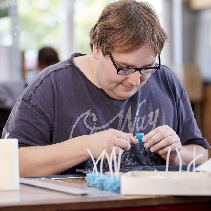 Ein Mann mit dunkelblauem T-Shirt und Brille fertigt Fädelkerzen an einem Tisch an. (vergrößerte Bildansicht wird geöffnet)