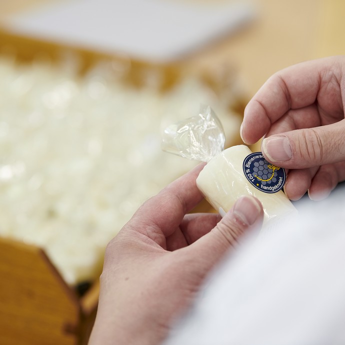 Männerhände kleben ein Etikett auf eine in Kunststofffolie verpackte Stumpenkerze. (vergrößerte Bildansicht wird geöffnet)